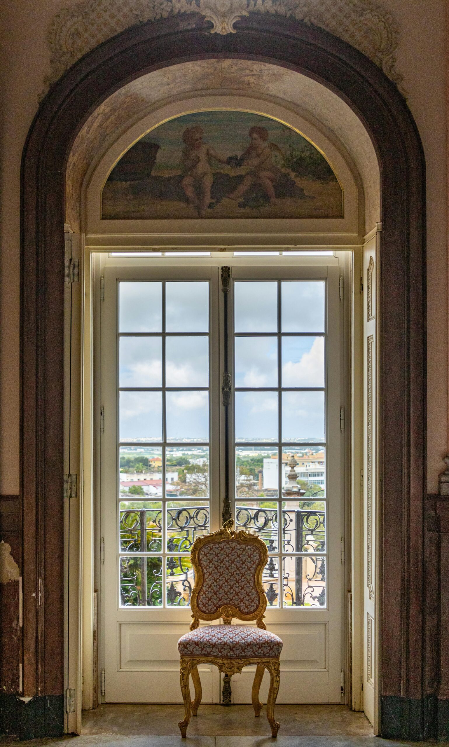 An ornate room with a classic chair facing a balcony, offering a scenic view of Estoi, Portugal.